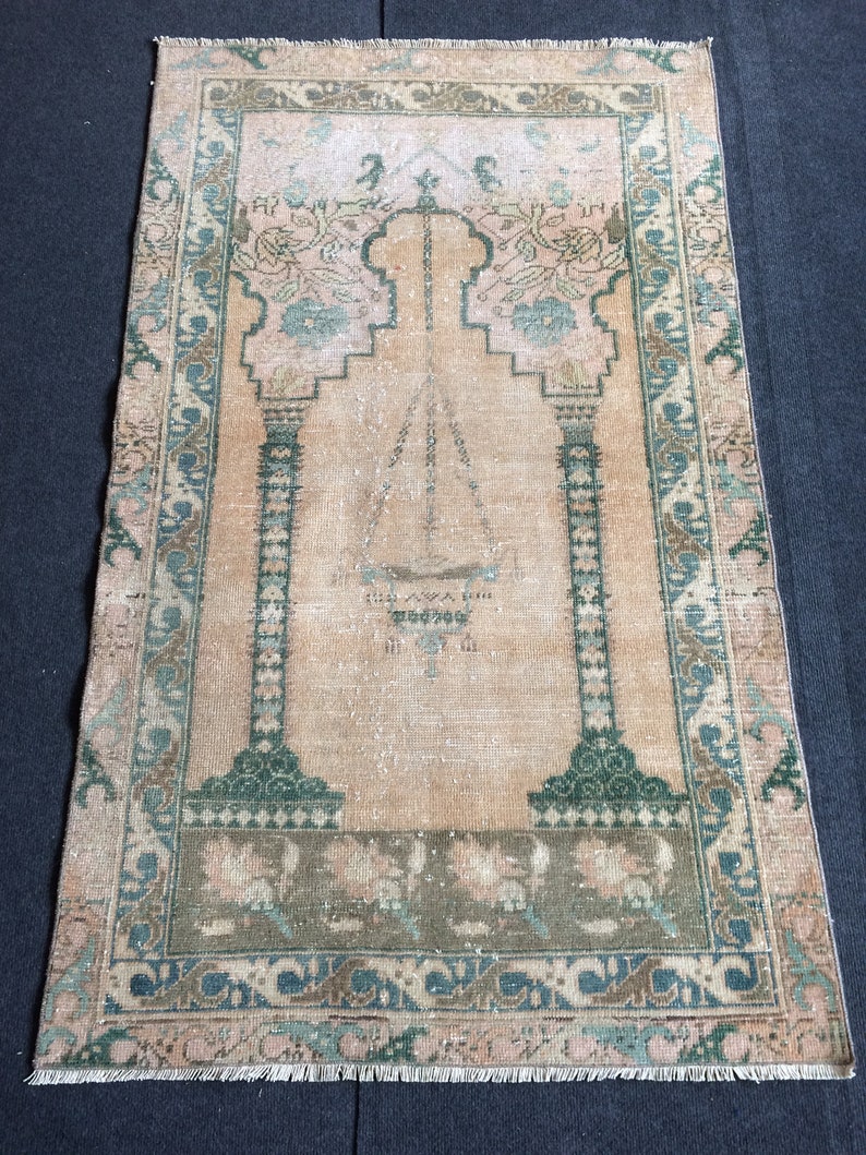 May include: A rectangular, vintage prayer rug with a central arch design. The rug features a peach-colored field with green and blue floral accents. The border is a mix of green, blue, and brown patterns, with a fringed edge.