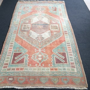 Puede incluir: Una alfombra rectangular de estilo vintage con un dise&ntilde;o geom&eacute;trico central en tonos coral, turquesa y crema. La alfombra presenta un borde detallado y una apariencia desgastada, lo que sugiere que es antigua o tiene un aspecto usado.