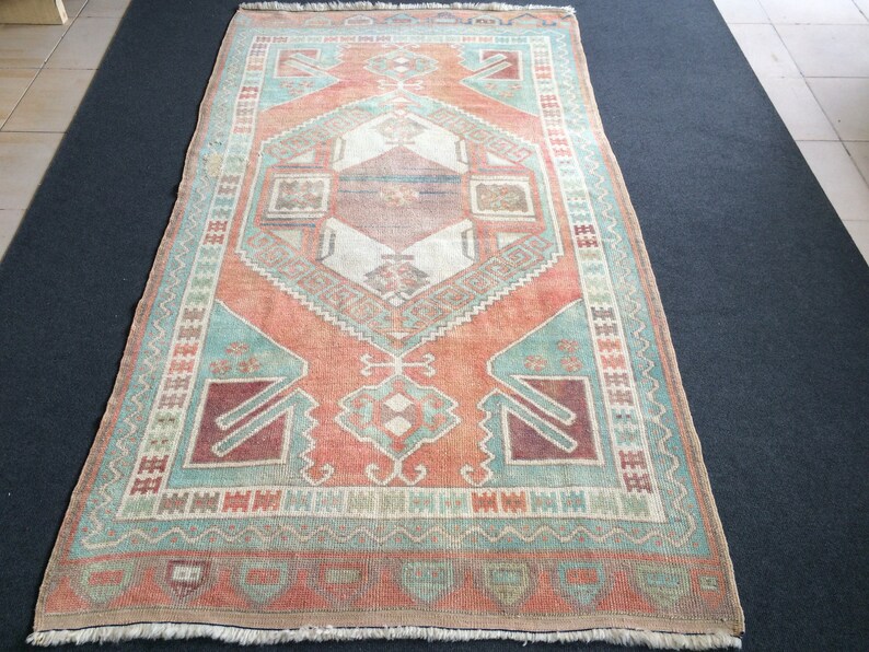 Puede incluir: Una alfombra rectangular de estilo vintage con un dise&ntilde;o geom&eacute;trico central en tonos coral, turquesa y crema. La alfombra presenta un borde detallado y una apariencia desgastada, lo que sugiere que es antigua o tiene un aspecto usado.