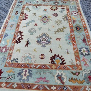 May include: A rectangular area rug with a cream-colored field and a decorative border. The rug features a repeating pattern of floral and abstract shapes in shades of blue, red, orange, and yellow. The border is a mix of blue, orange, and cream.