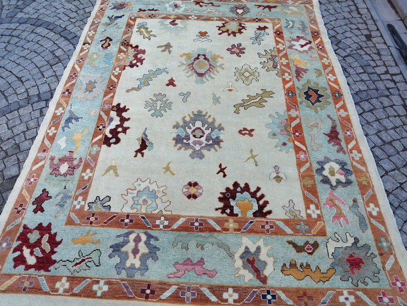 May include: A rectangular area rug with a cream-colored field and a decorative border. The rug features a repeating pattern of floral and abstract shapes in shades of blue, red, orange, and yellow. The border is a mix of blue, orange, and cream.