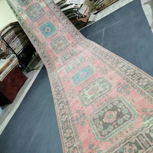 May include: A long, rectangular rug with a faded, vintage appearance. The rug features a pink central field with repeating geometric patterns in teal, gray, and cream. The border is a darker gray with floral motifs. The rug is likely made of woven material.