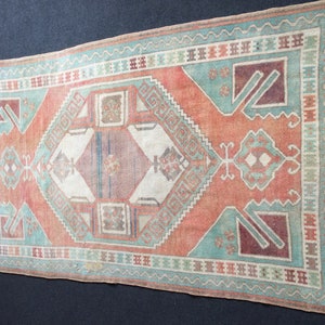 Puede incluir: Alfombra rectangular de estilo vintage con aspecto desgastado. Presenta un dise&ntilde;o central de rombos en tonos melocot&oacute;n, turquesa y burdeos, enmarcado por bordes geom&eacute;tricos. Flecos en los extremos cortos acent&uacute;an su est&eacute;tica envejecida.