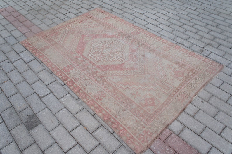 May include: A rectangular, distressed rug with a central geometric design and a border. The rug features a faded, vintage aesthetic with a color palette of beige and muted red. The rug is placed on a brick surface.