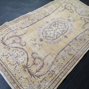 Puede incluir: Una alfombra rectangular de estilo vintage con un fondo beige y intrincados patrones florales y de vid en tonos marrones y blancos. La alfombra tiene un borde detallado y un diseño central de medallón, creando una apariencia clásica y envejecida.