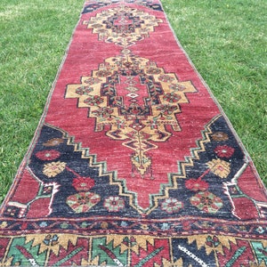 Puede incluir: Una alfombra roja con un intrincado diseño geométrico. La alfombra presenta un diseño central en dorado y negro, con detalles en verde y azul. La alfombra está colocada sobre hierba verde.