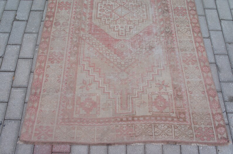 May include: A rectangular, distressed rug with a faded, vintage appearance. The rug features a central geometric design in shades of pink and beige, framed by a detailed border. The rug's design includes floral and geometric patterns.