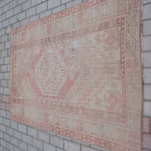 May include: A rectangular, vintage rug with a faded, distressed appearance. The rug features a central diamond pattern in shades of pink and beige, surrounded by geometric borders. The rug is displayed against a gray brick wall.