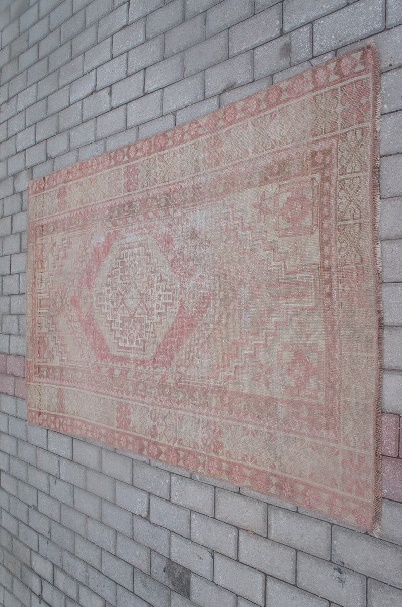 May include: A rectangular, vintage rug with a faded, distressed appearance. The rug features a central diamond pattern in shades of pink and beige, surrounded by geometric borders. The rug is displayed against a gray brick wall.