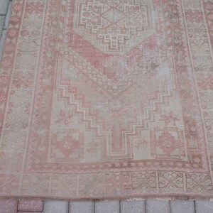 May include: A rectangular, distressed rug with a faded, vintage appearance. The rug features a central geometric design in shades of pink and beige, framed by a detailed border. The rug's design includes floral and geometric patterns.