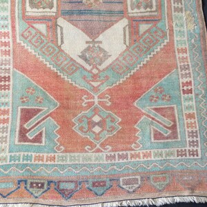 Puede incluir: Una alfombra rectangular de estilo vintage con un dise&ntilde;o central en tonos melocot&oacute;n, turquesa y burdeos. La alfombra presenta patrones geom&eacute;tricos y un borde con flecos. El dise&ntilde;o general es sim&eacute;trico, con una apariencia desgastada.