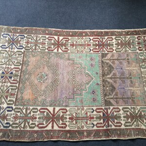 May include: Rectangular vintage rug with a distressed look. The rug has a central design in shades of purple, green, and brown, framed by a border with geometric patterns in red, blue, and beige. The rug's design is symmetrical.
