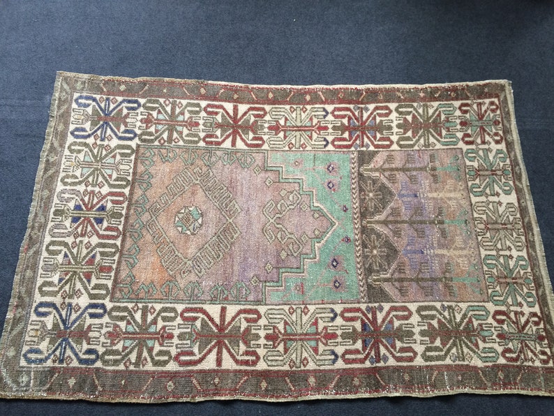 May include: Rectangular vintage rug with a distressed look. The rug has a central design in shades of purple, green, and brown, framed by a border with geometric patterns in red, blue, and beige. The rug's design is symmetrical.