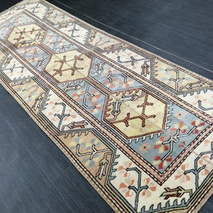 Könnte beinhalten: Ein rechteckiger Teppich mit einem zentralen geometrischen Muster in Beige, Hellblau und Rostfarben. Der Teppich hat einen Rand mit sich wiederholenden floralen und geometrischen Motiven. Der Teppich ist wahrscheinlich aus Wolle oder einem ähnlichen Material.