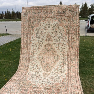 Puede incluir: Una alfombra rectangular grande con un intrincado diseño floral. La alfombra presenta un fondo color crema con un medallón central y un borde en tonos melocotón y marrón. La alfombra se exhibe al aire libre sobre una zona de césped.