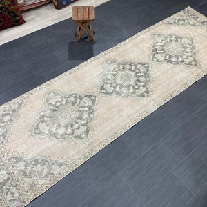 May include: A long, rectangular rug with a beige background and intricate gray floral patterns. The rug features a border design and three large central medallions. The rug is placed on a dark tiled floor.