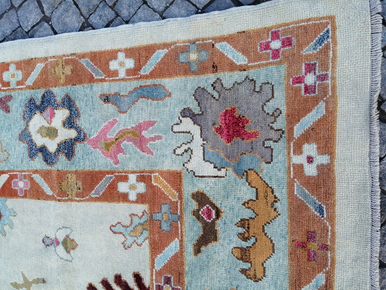 May include: Close-up of a section of a patterned rug. The rug features a light blue central field with floral and geometric designs in shades of brown, pink, and gray. The border is a darker brown with repeating floral motifs.