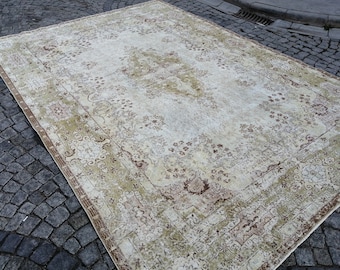 May include: A large, rectangular, vintage-style rug with an intricate floral pattern. The rug features a cream-colored central field with brown and green floral motifs and a detailed border. The rug is placed on a cobblestone surface.
