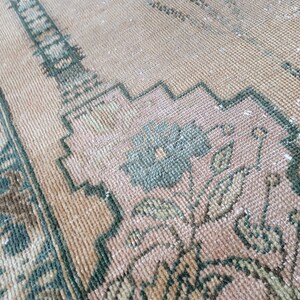 May include: Close-up of a vintage rug with a faded, distressed appearance. The rug features a peach-colored background with intricate floral and geometric patterns in shades of green and brown. The texture appears soft and worn.