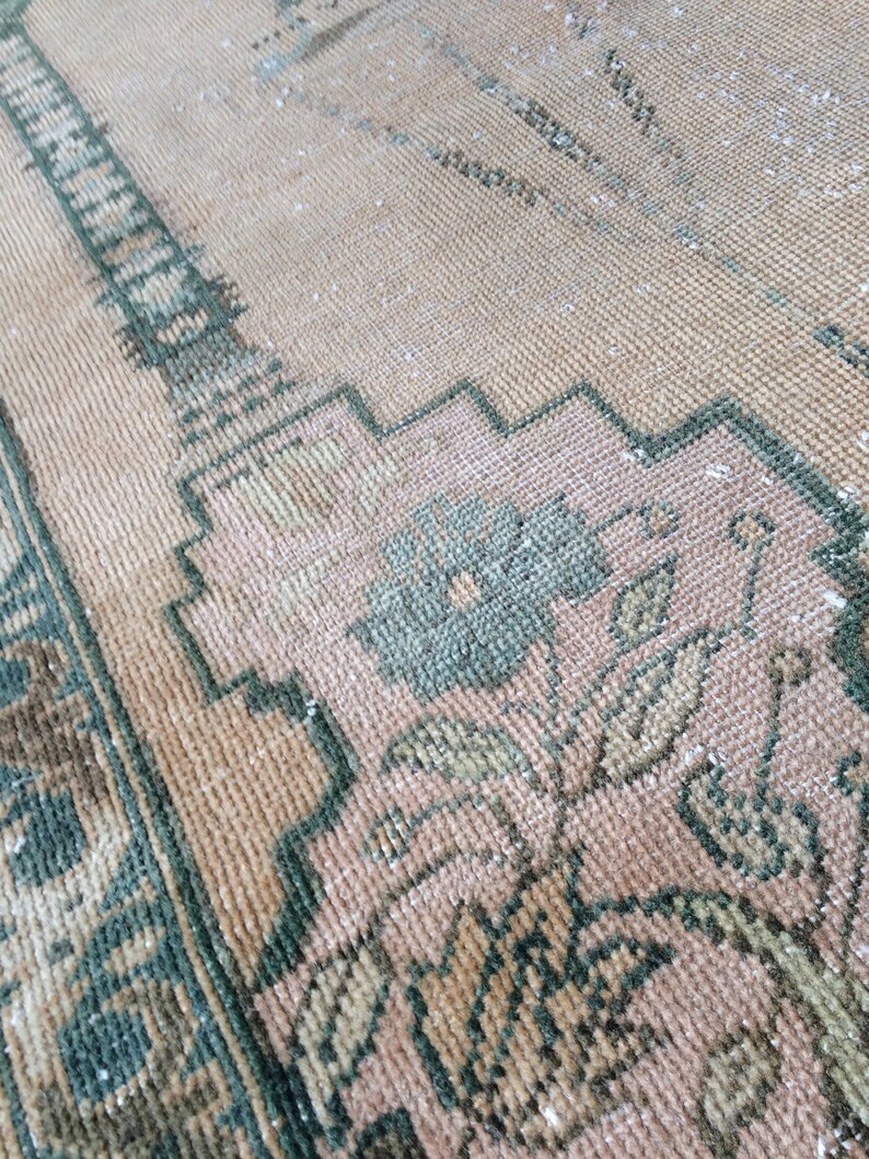 May include: Close-up of a vintage rug with a faded, distressed appearance. The rug features a peach-colored background with intricate floral and geometric patterns in shades of green and brown. The texture appears soft and worn.