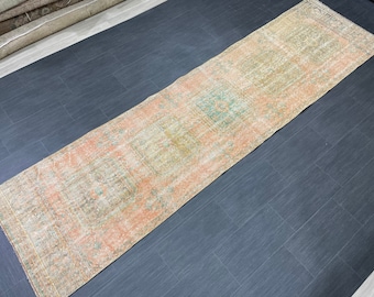 May include: A long, rectangular rug with a distressed, vintage appearance. The rug features a central design with faded peach, green, and beige tones. The rug's pattern includes geometric shapes and borders, suitable for a hallway or entryway.