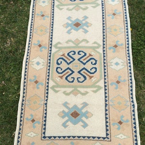 May include: A rectangular, beige-colored rug with a central cream-colored panel. The rug features geometric patterns in blue, brown, and green. The border is a lighter beige with repeating geometric shapes. The rug has a fringe on both ends.