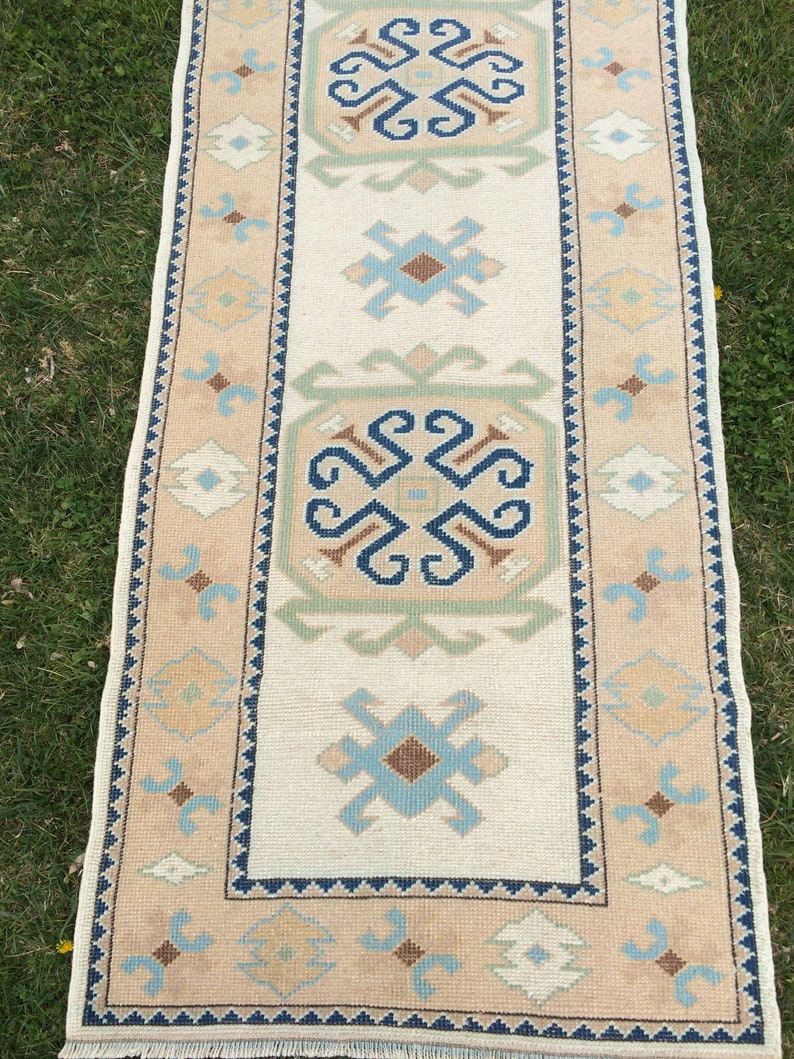 May include: A rectangular, beige-colored rug with a central cream-colored panel. The rug features geometric patterns in blue, brown, and green. The border is a lighter beige with repeating geometric shapes. The rug has a fringe on both ends.