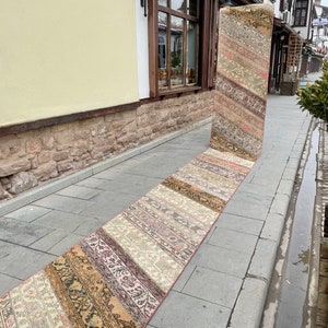 May include: A long, narrow, patchwork rug with a variety of colors and patterns. The rug is laid out on a sidewalk, showcasing its length and design. The rug features a mix of beige, brown, pink, and green tones, creating a vintage aesthetic.