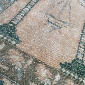 May include: A vintage rug with a distressed appearance. The rug features a central field in a warm beige hue, framed by borders in shades of green, brown, and hints of pink. The design includes geometric and floral patterns, creating a classic, aged aesthetic.
