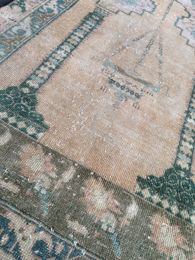 May include: A vintage rug with a distressed appearance. The rug features a central field in a warm beige hue, framed by borders in shades of green, brown, and hints of pink. The design includes geometric and floral patterns, creating a classic, aged aesthetic.