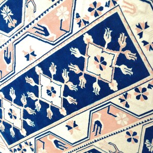 May include: Close-up of a patterned rug with a geometric design. The rug features a navy blue central panel with white and pink accents. Floral and stylized hand motifs are incorporated into the design.