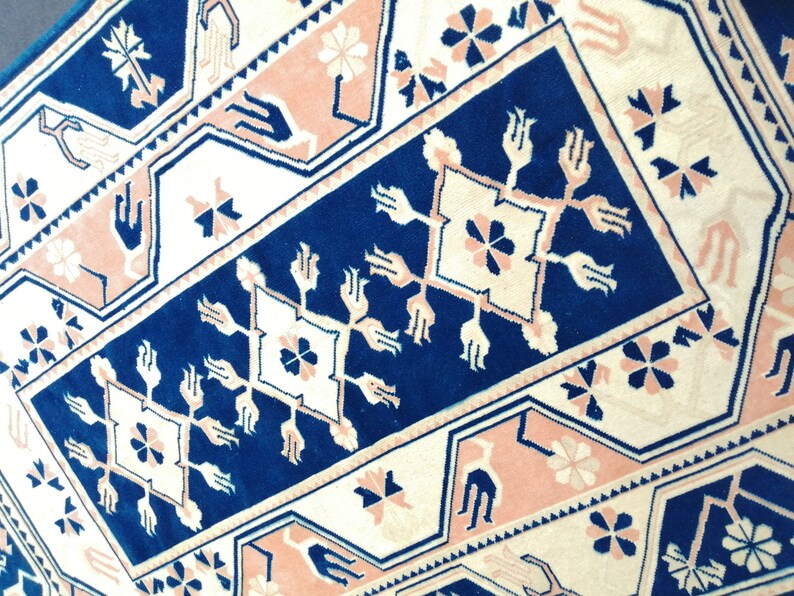 May include: Close-up of a patterned rug with a geometric design. The rug features a navy blue central panel with white and pink accents. Floral and stylized hand motifs are incorporated into the design.