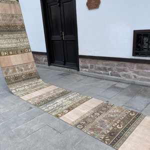 May include: A long, rectangular runner rug with a patchwork design. The rug features alternating stripes of beige and patterned sections in shades of green, brown, and cream. The rug is displayed outdoors, leaning against a white wall with a black door.