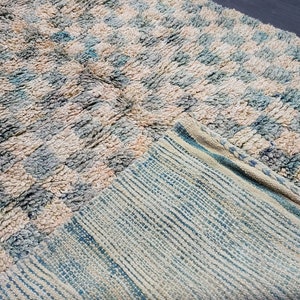 Op de afbeelding: Een getextureerd tapijt met een geruit patroon in cr&egrave;me, lichtblauw en beige tinten. De pool van het tapijt is zichtbaar en een deel van de onderkant, met een gestreept ontwerp, is zichtbaar. Het tapijt is opgevouwen en onthult de contrasterende texturen en kleuren.