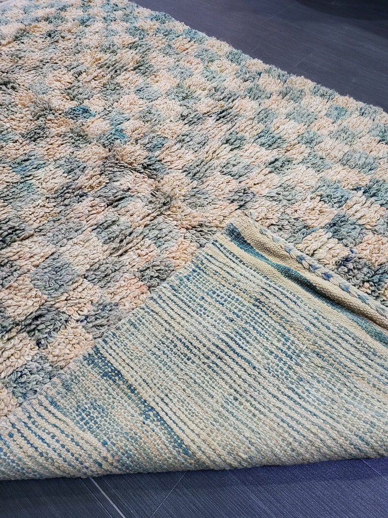 Op de afbeelding: Een getextureerd tapijt met een geruit patroon in cr&egrave;me, lichtblauw en beige tinten. De pool van het tapijt is zichtbaar en een deel van de onderkant, met een gestreept ontwerp, is zichtbaar. Het tapijt is opgevouwen en onthult de contrasterende texturen en kleuren.