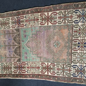 May include: A rectangular vintage rug with a central design in shades of purple, green, and orange. The rug has a border with geometric patterns in brown, red, and blue. The rug's aged appearance adds to its charm.
