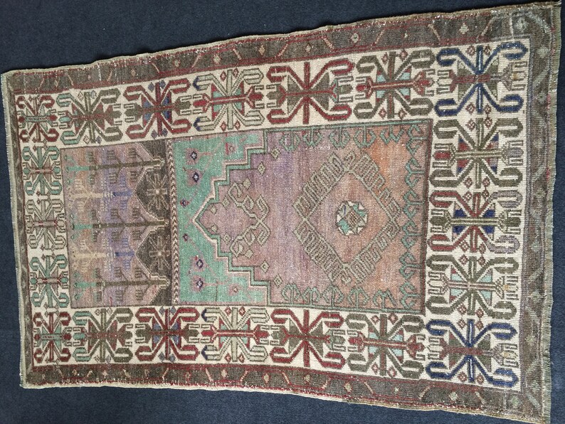 May include: A rectangular vintage rug with a central design in shades of purple, green, and orange. The rug has a border with geometric patterns in brown, red, and blue. The rug's aged appearance adds to its charm.
