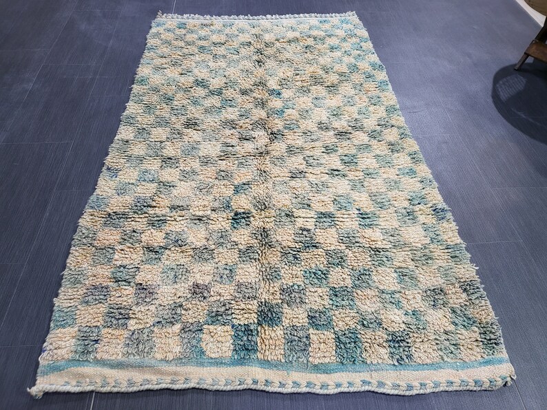 Op de afbeelding: Rechthoekig vloerkleed met een geblokt patroon in beige en turquoise. Het kleed heeft een getextureerde, geweven uitstraling en een lichtere rand. Geschikt voor een woon- of slaapkamer, meet ongeveer 150 cm x 250 cm.