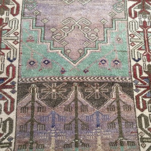 May include: A rectangular vintage rug with a faded, distressed look. The rug's central design features lavender, teal, and brown hues, with geometric patterns. The border is in red, brown, and cream. The rug's texture suggests it is made of woven material.