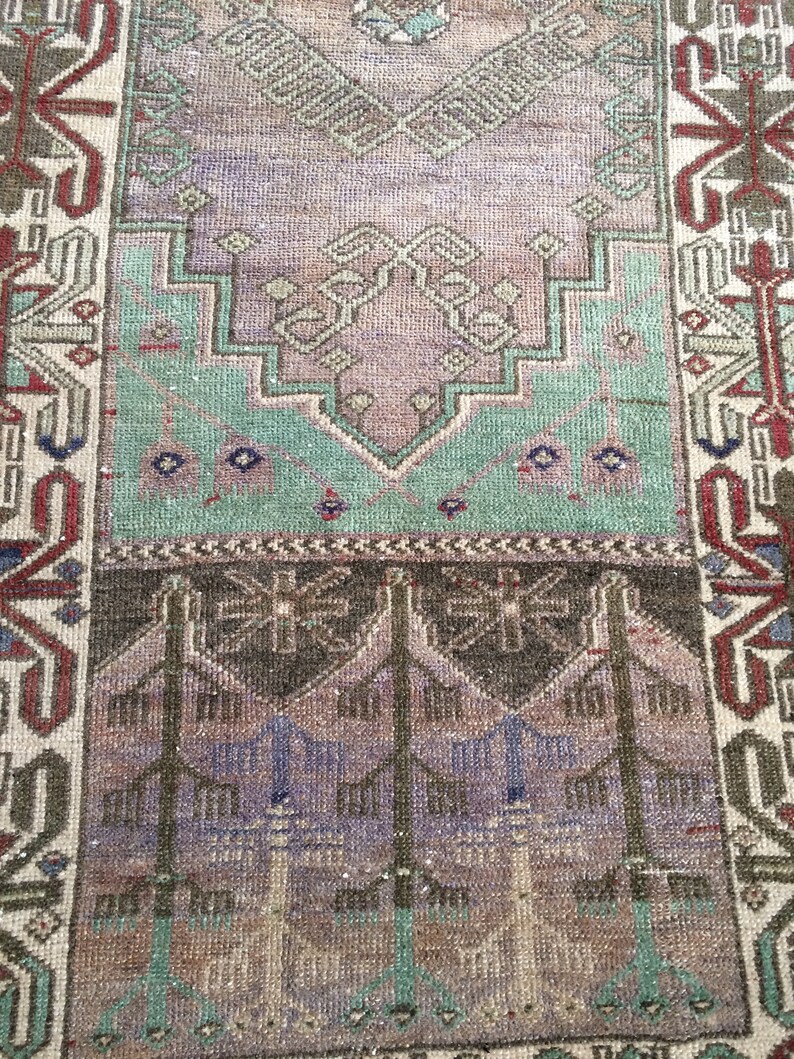 May include: A rectangular vintage rug with a faded, distressed look. The rug's central design features lavender, teal, and brown hues, with geometric patterns. The border is in red, brown, and cream. The rug's texture suggests it is made of woven material.