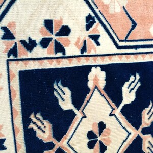 May include: Close-up of a section of a rug with a geometric pattern. The rug features a cream-colored background with navy blue and peach-colored geometric shapes and floral designs. The border is navy blue.