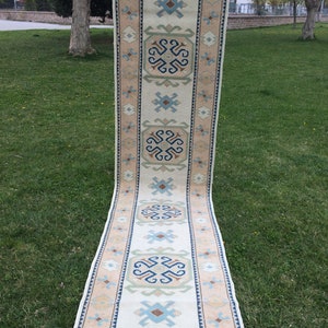 May include: A long, rectangular rug with a cream-colored central field and a border in shades of tan, blue, and green. The rug features geometric patterns and a fringed edge. The rug is displayed outdoors on a grassy surface.
