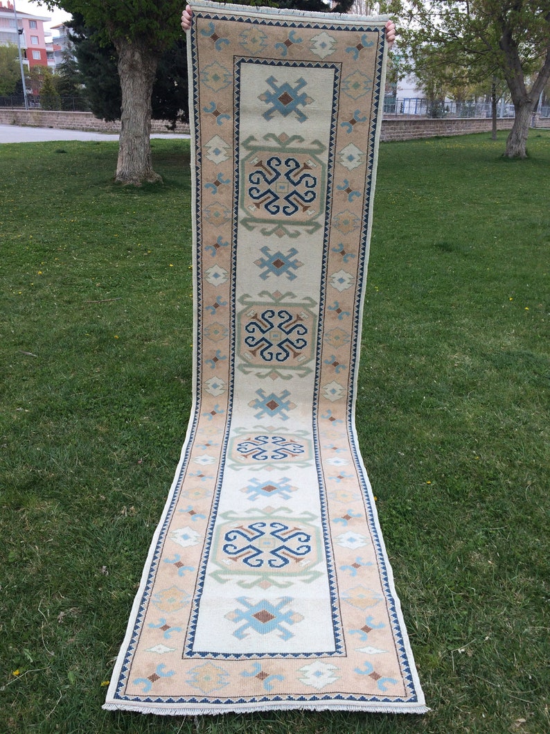 May include: A long, rectangular rug with a cream-colored central field and a border in shades of tan, blue, and green. The rug features geometric patterns and a fringed edge. The rug is displayed outdoors on a grassy surface.