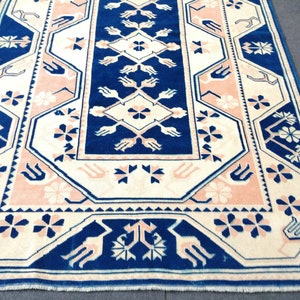 May include: A rectangular rug with a central navy blue panel featuring a diamond and floral design. The rug has a cream-colored border with pink and navy blue floral and geometric patterns. The rug's dimensions are not visible.