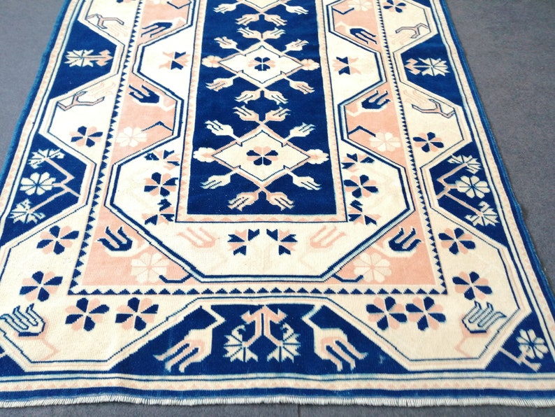 May include: A rectangular rug with a central navy blue panel featuring a diamond and floral design. The rug has a cream-colored border with pink and navy blue floral and geometric patterns. The rug's dimensions are not visible.