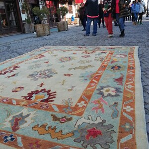 May include: A large, rectangular rug with a cream-colored central field and a border in shades of teal, orange, and burgundy. The rug features floral and abstract patterns. The rug is displayed outdoors on a cobblestone street, with people and buildings in the background.