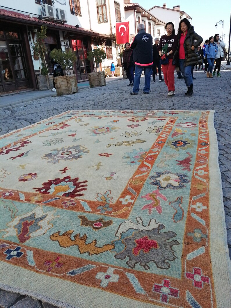 May include: A large, rectangular rug with a cream-colored central field and a border in shades of teal, orange, and burgundy. The rug features floral and abstract patterns. The rug is displayed outdoors on a cobblestone street, with people and buildings in the background.