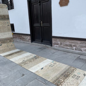 May include: A long, rectangular runner rug with a patchwork design. The rug features a variety of neutral tones, including beige, cream, and brown, with geometric and floral patterns. The rug is displayed outdoors on a stone surface.