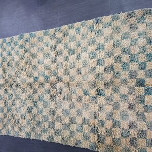 Op de afbeelding: Een rechthoekig tapijt met een geblokt patroon in cr&egrave;me en blauwgroen. Het tapijt heeft een getextureerde, geweven uitstraling en een cr&egrave;mekleurige rand met een blauwgroene rand. Het tapijt is geschikt voor woondecoratie.
