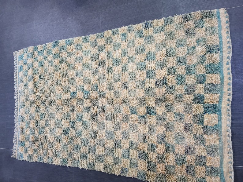 Op de afbeelding: Een rechthoekig tapijt met een geblokt patroon in cr&egrave;me en blauwgroen. Het tapijt heeft een getextureerde, geweven uitstraling en een cr&egrave;mekleurige rand met een blauwgroene rand. Het tapijt is geschikt voor woondecoratie.